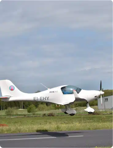 A small white aeroplane with the registration EI-EHY is taking off or landing on a runway surrounded by green grass and trees under a partly cloudy sky.