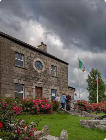 A stone building with a clock above the door and two flags outside, one Irish and one French. Three people stand talking near the entrance, and pink flowers bloom in the garden. The sky is cloudy and overcast.