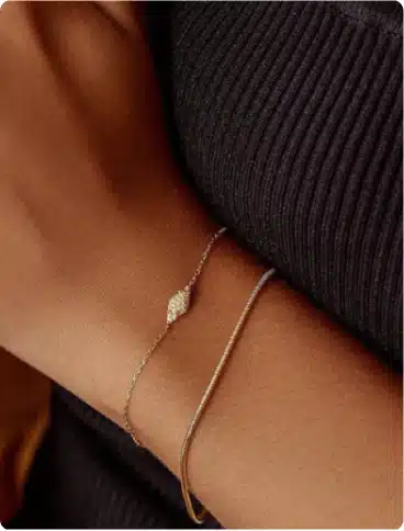 A close-up of a person's wrist wearing two delicate bracelets, one with a small diamond-shaped charm and the other a simple gold band, against a dark ribbed fabric background.