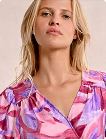A woman with long blonde hair wears a V-neck dress featuring a pink, purple, and red abstract pattern. She is looking confidently at the camera against a plain, light-coloured background.