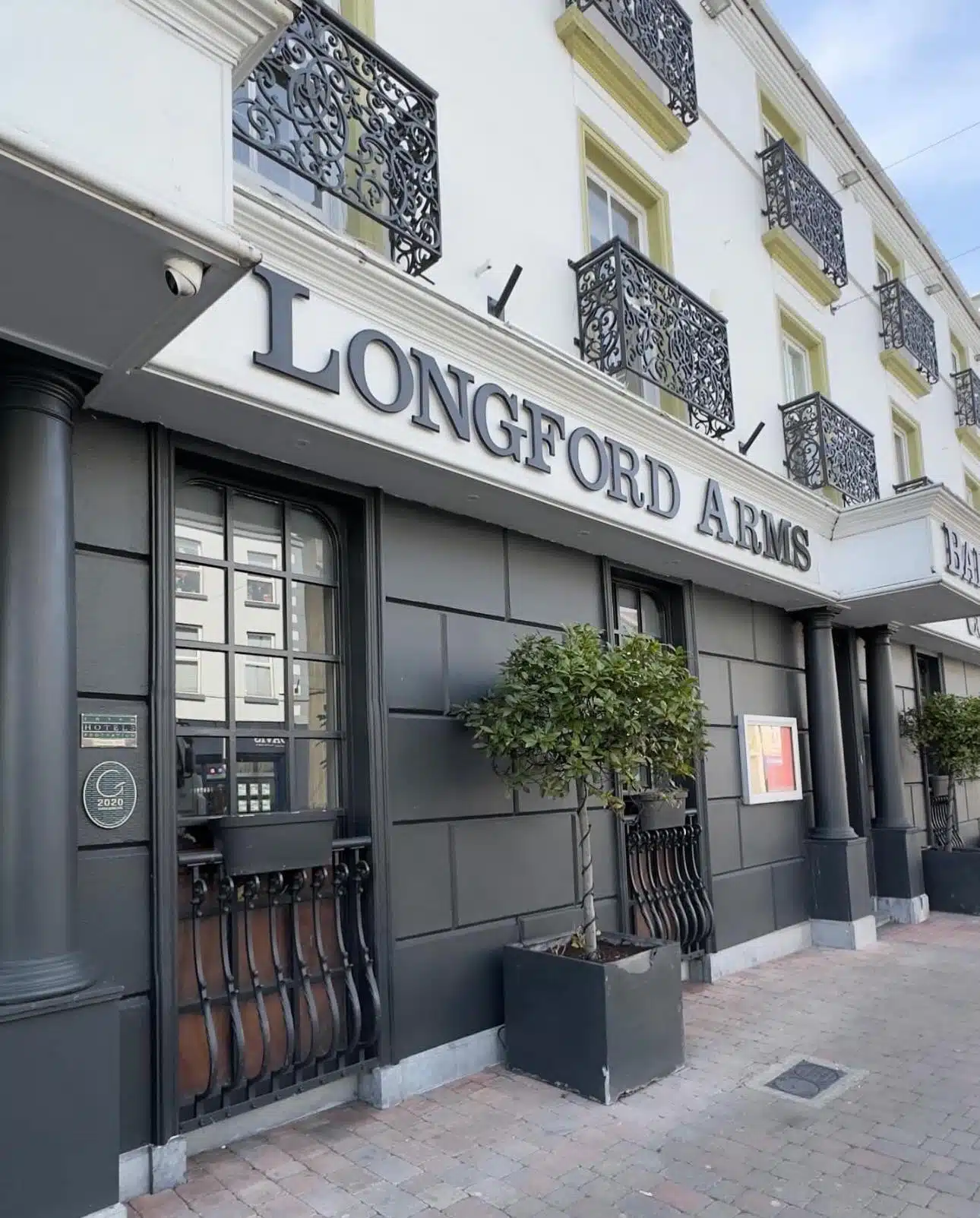 A hotel or pub exterior with a sign reading "LONGFORD ARMS," featuring decorative black railings, potted trees, and ornate balcony railings on the upper storeys.