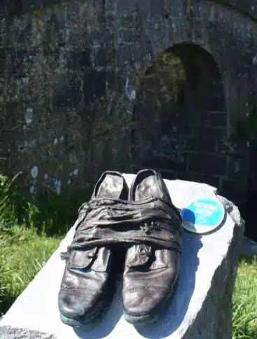 A pair of worn, dark shoes wrapped with cloth sit on a stone slab outdoors, with an arched stone bridge and grass in the blurred background. A round blue badge is next to the shoes.